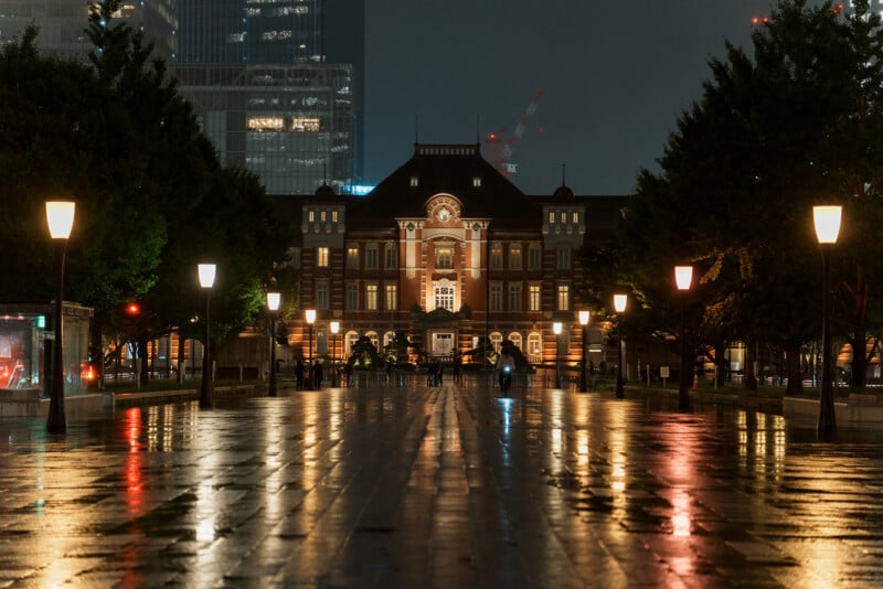 Tokyo Station at night, illuminated by streetlights with reflections on the wet pavement, surrounded by trees and modern buildings in the background.