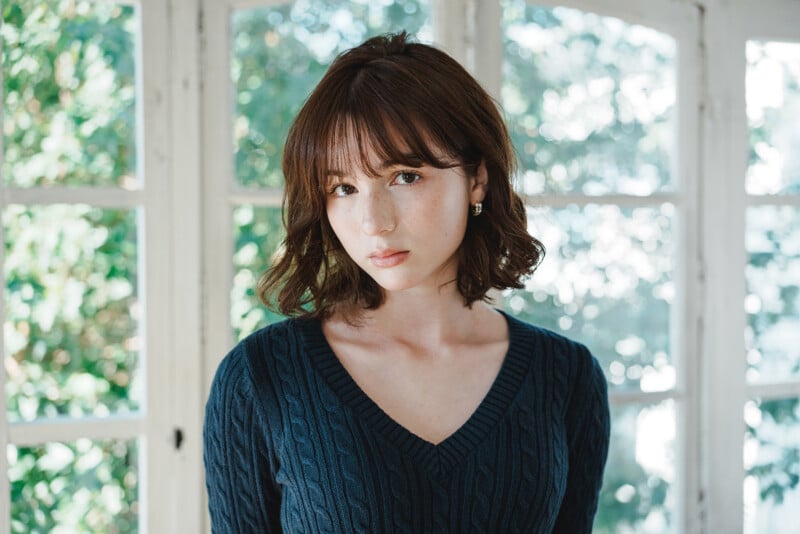 A young woman with short brown hair and bangs stands indoors in front of large windows, wearing a dark blue knit sweater. Sunlight filters through green foliage outside, creating a soft, natural background.