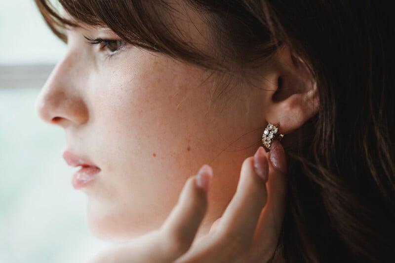 A close-up of a woman with fair skin touching her ear, wearing a sparkling, gem-studded earring. Her brown hair partially covers her face, and her expression is calm and thoughtful.