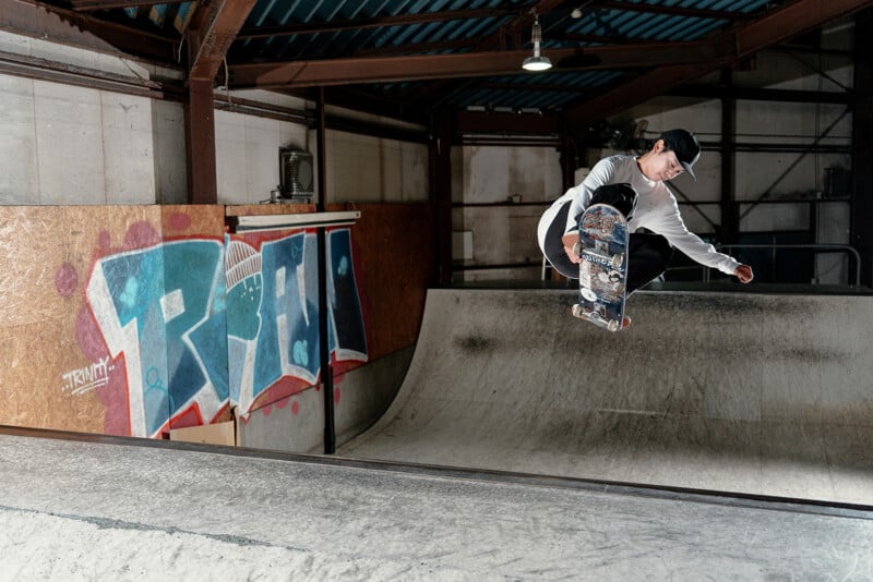 A skateboarder in a white shirt and black cap performs an aerial trick above an indoor skate ramp, with colorful graffiti art visible on the wall behind him.