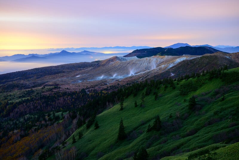 Sunrise over a mountainous landscape with green hills in the foreground, a steaming crater in the midground, and layers of distant blue mountains under a colorful sky.