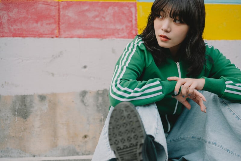 A young person with wavy dark hair, wearing a green jacket with white stripes and light blue jeans, sits on the ground in front of a colorful wall, looking to the side with a thoughtful expression.