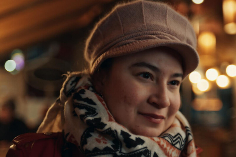 A woman wearing a pink hat and patterned scarf smiles softly, with warm, blurred lights and a cozy indoor setting in the background.