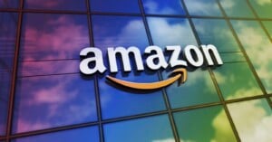 The Amazon logo with a yellow arrow is displayed on the glass exterior of a building, reflecting a colorful, cloudy sky.