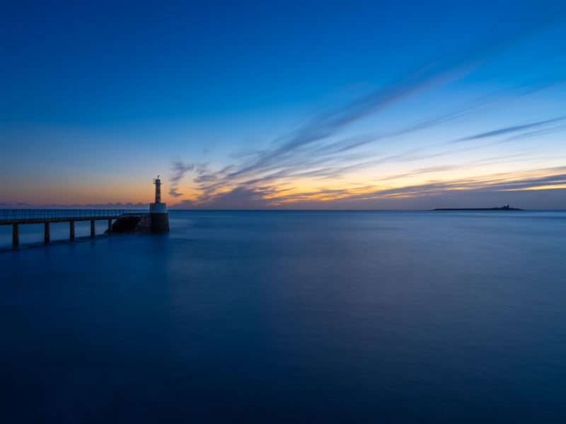 A lighthouse stands at the end of a pier overlooking calm, blue water at sunset, with soft clouds streaking across the colorful sky.