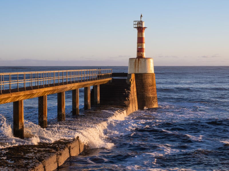 A red and white striped lighthouse stands at the end of a pier, with waves crashing against the structure, under a clear sky at sunset.