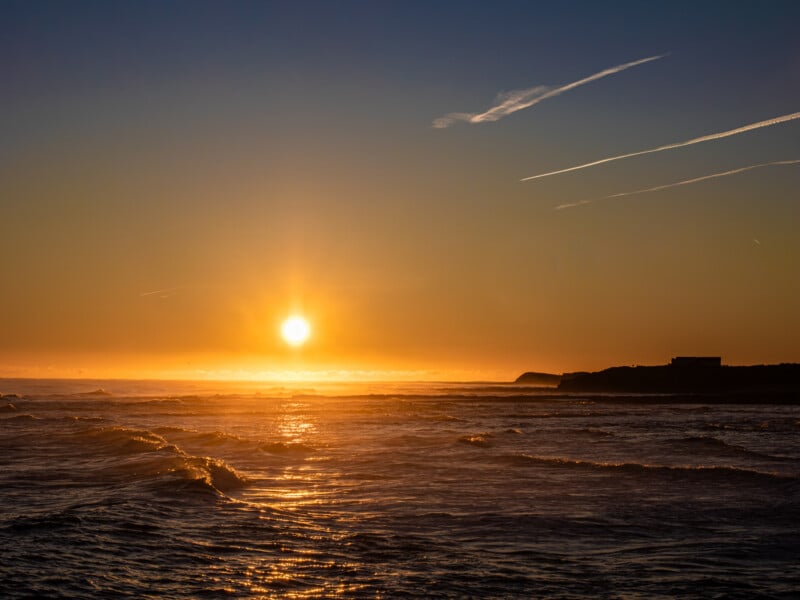 Golden sun setting over a calm ocean with gentle waves, orange light reflecting on the water, and a silhouetted coastline under a clear sky with streaks of clouds.