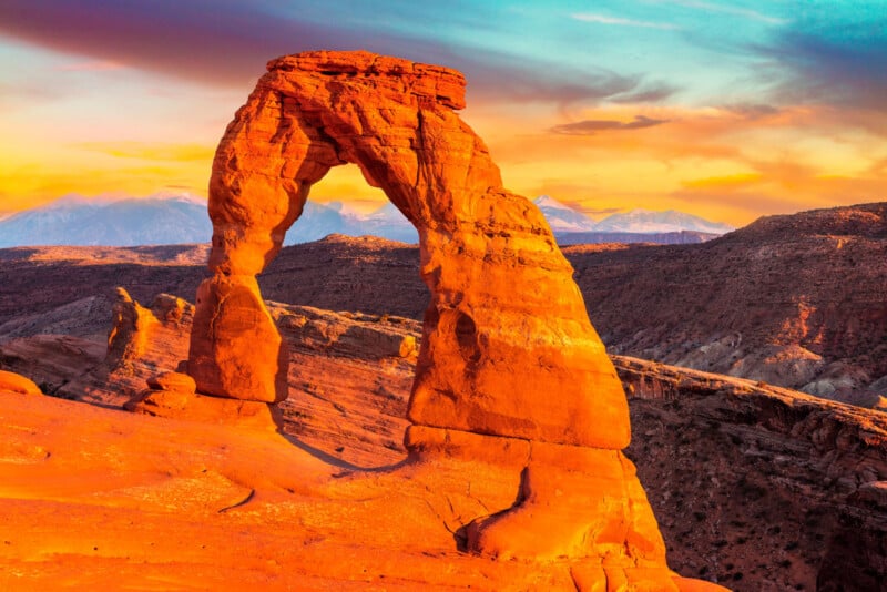 A natural sandstone arch, Delicate Arch, stands in a desert landscape in Arches National Park, Utah, illuminated by warm sunset light. Distant mountains and a colorful sky are visible in the background.