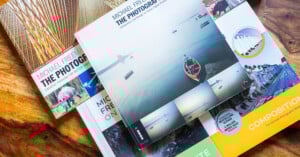 A collection of photography books by Michael Freeman are spread out on a wooden surface, with colorful covers featuring images of landscapes, animals, and people. The central book displays a serene water scene with a red boat.