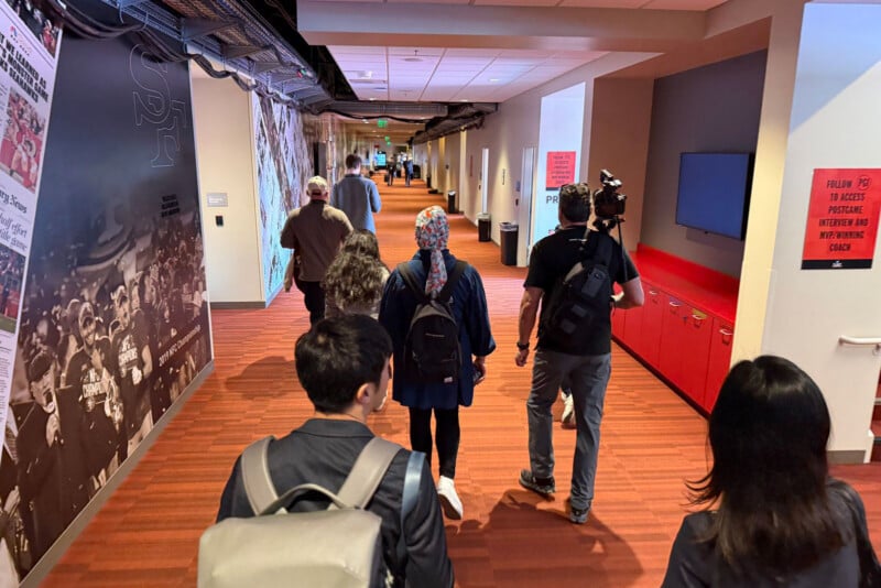 A group of people walk down a brightly lit hallway with red carpet and walls, some carrying backpacks. One person has a camera on their shoulder. Posters and signage are visible on the walls.