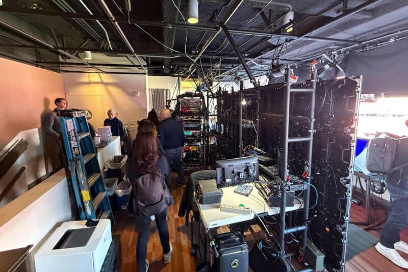 People walk through a busy backstage area filled with computer equipment, cables, and monitors. Various technical gear is set up on shelves and tables; some individuals are working while others move through the space.