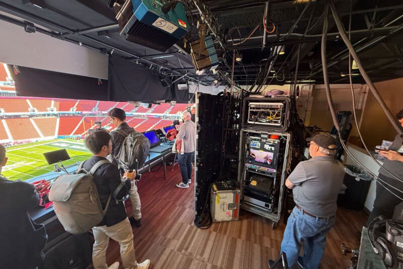 A broadcast booth overlooks a football stadium, with several people standing near technical equipment and monitors; some are working while others observe the field below.