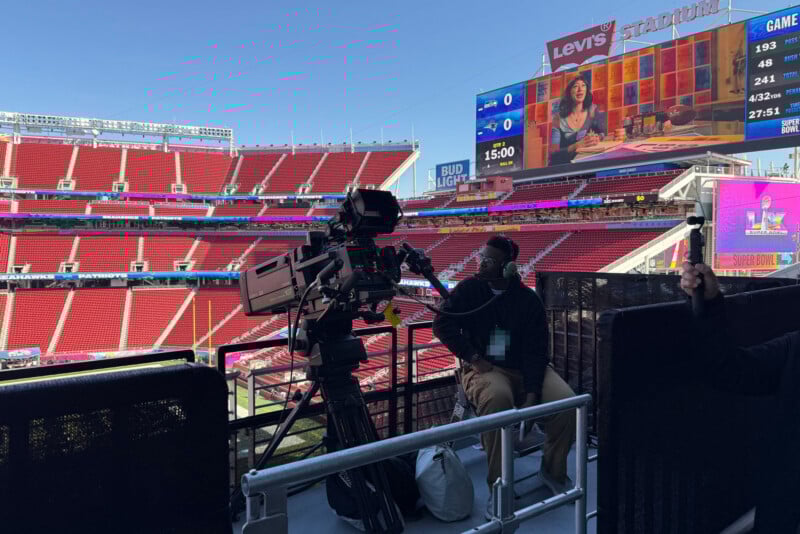 A cameraman in a mask operates a large TV camera at an empty stadium with red seats. A scoreboard and a large screen showing a woman talking are visible in the background. It's a bright, sunny day at Levi’s Stadium.