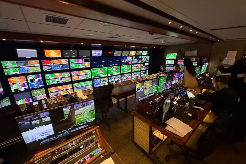 A busy broadcast control room with numerous monitors displaying sports events, graphics, and color bars. Several people are seated at desks, operating equipment and managing the live broadcast.