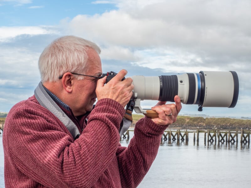 Un hombre con cabello gris y gafas se encuentra cerca de una masa de agua con un muelle de madera al fondo, usando una cámara OM-1 Mark II equipada con una lente OM System 150-400 F4.5 PRO. Con una chaqueta granate, miró por el visor de su cámara.