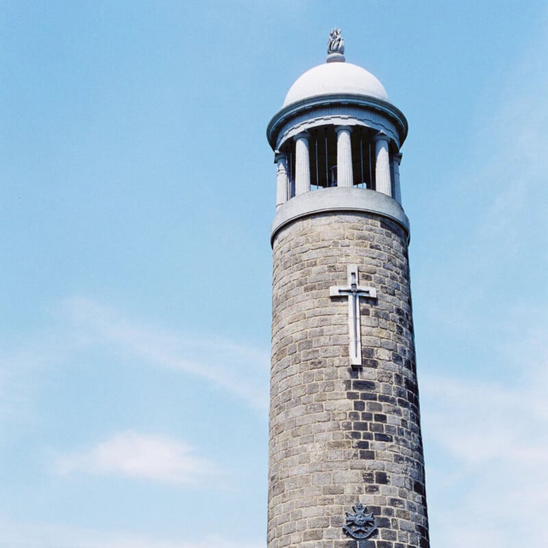 A tall cylindrical stone tower with a white domed roof and open pillars, featuring a large cross mounted on its front, set against a clear blue sky.