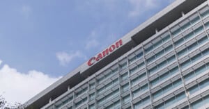 A modern glass office building with the red Canon logo on the upper facade, set against a blue sky with some clouds.