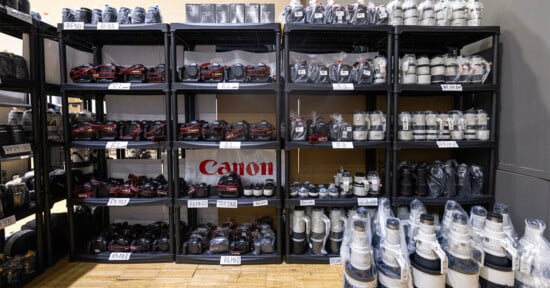 Shelves filled with Canon cameras and various camera lenses, all neatly arranged and wrapped in plastic, with a Canon logo banner displayed in the center.