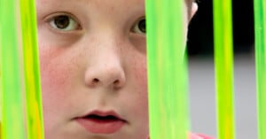 A close-up of a child's face with freckles, wide eyes, and parted lips, looking through vertical neon green translucent bars.