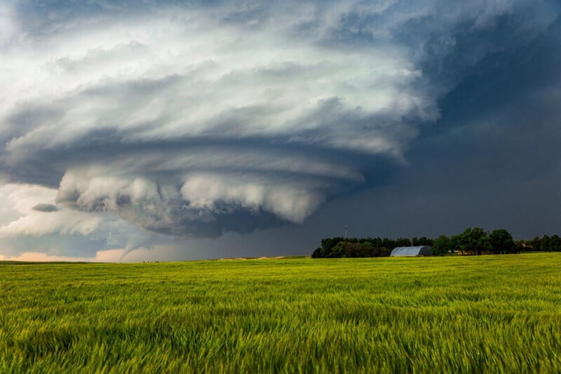 Una espectacular súper tormenta y nubes arremolinadas se ciernen sobre un campo verde, con un pequeño granero y árboles visibles en la distancia bajo un cielo oscuro y tormentoso.