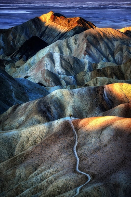 Un sendero sinuoso atraviesa colinas coloridas e iluminadas por el sol, con un excursionista solitario en el medio. El paisaje ondulado presenta sombras y crestas espectaculares contra un cielo vívido.