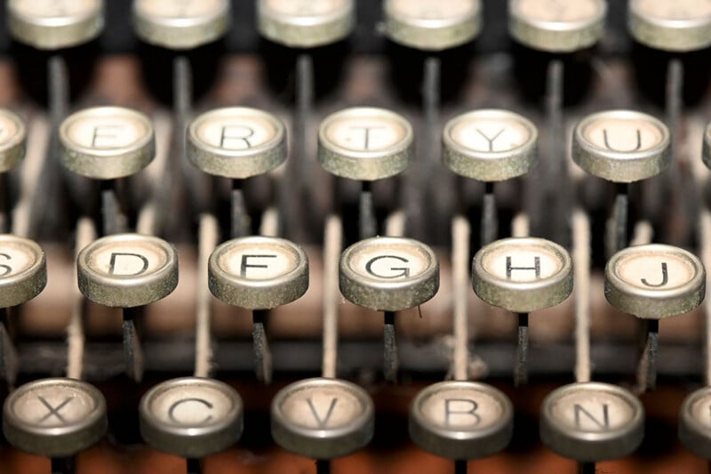 Primer plano de teclas de máquinas de escribir antiguas que muestran botones redondos desgastados con letras negras sobre fondo blanco y varillas de metal que los mantienen en su lugar. Las teclas están dispuestas en ordenadas filas horizontales.