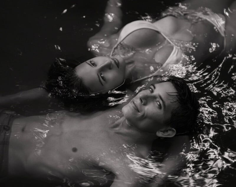 Un hombre y una mujer flotan boca arriba en el agua, ambos mirando hacia arriba. La mujer lleva bikini y el hombre está en topless. La imagen está en blanco y negro con suaves ondulaciones a su alrededor.