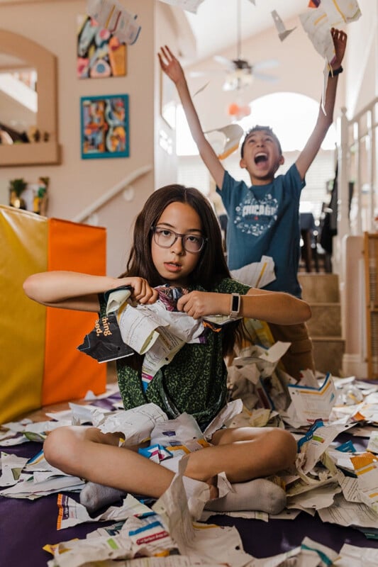 Una niña está sentada en el suelo con las piernas cruzadas, rompiendo papel con expresión seria, mientras un niño detrás de ella está de pie en las escaleras, con los brazos levantados con emoción. Un salón colorido con papeles rotos esparcidos por todas partes.