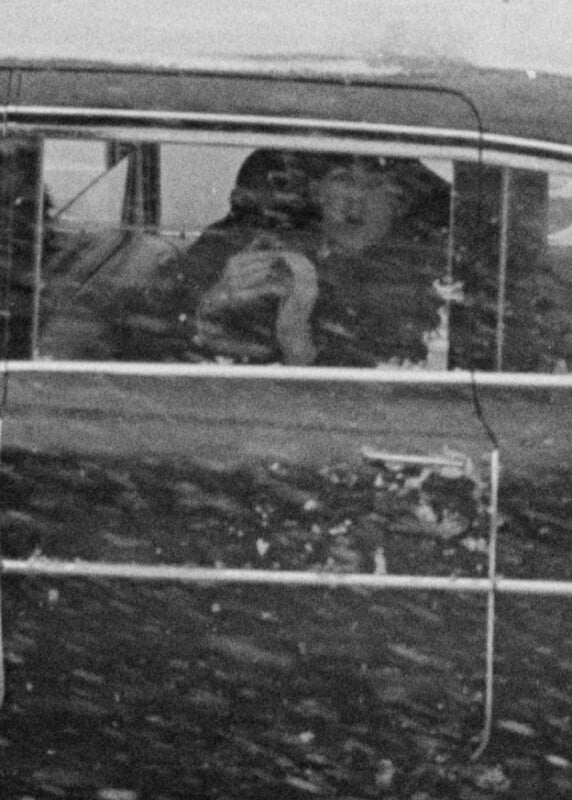 A black and white photo of a person sitting in the back seat of a car, partially obscured by the window. The person appears to be wearing a hood and holding something in their hands near their face.