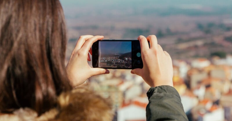 A person with long brown hair wearing a green jacket takes a photo of a scenic, sunlit landscape with their smartphone, capturing a distant town and natural surroundings.
