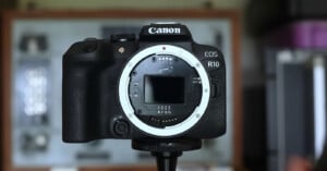A Canon EOS R10 digital camera body without a lens is mounted on a tripod, with a blurred background showing shelves and equipment.