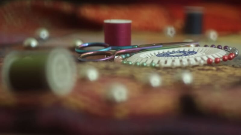 Primer plano de artículos de costura que incluyen tijeras, bobinas, portaalfileres redondos con alfileres de colores y botones dispersos sobre una superficie de tela texturizada. Tanto el primer plano como el fondo están borrosos.