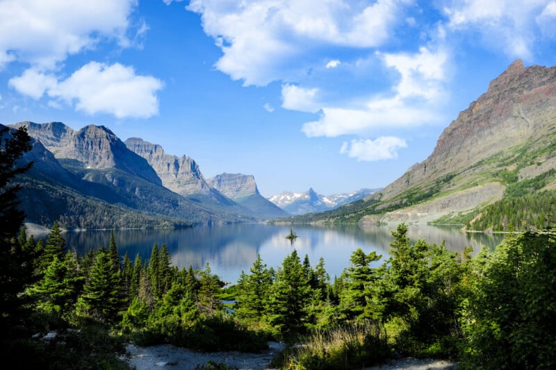 A scenic view of a calm lake surrounded by pine trees and tall mountains under a bright blue sky with scattered white clouds. Snow-capped peaks are visible in the distance.