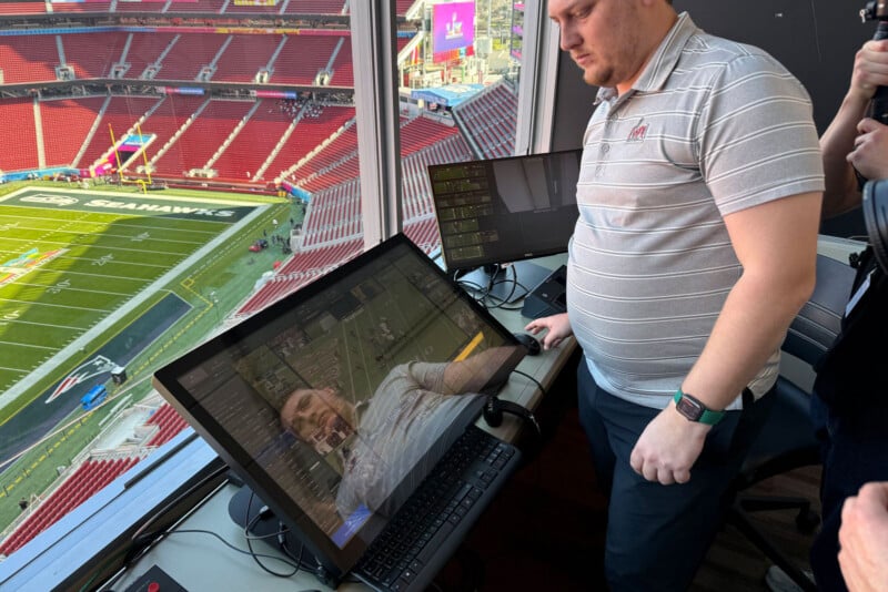 A man in a striped polo shirt stands by a large touchscreen monitor displaying a football field. He is in a stadium press box overlooking an empty field and stands, with another monitor and a person nearby.
