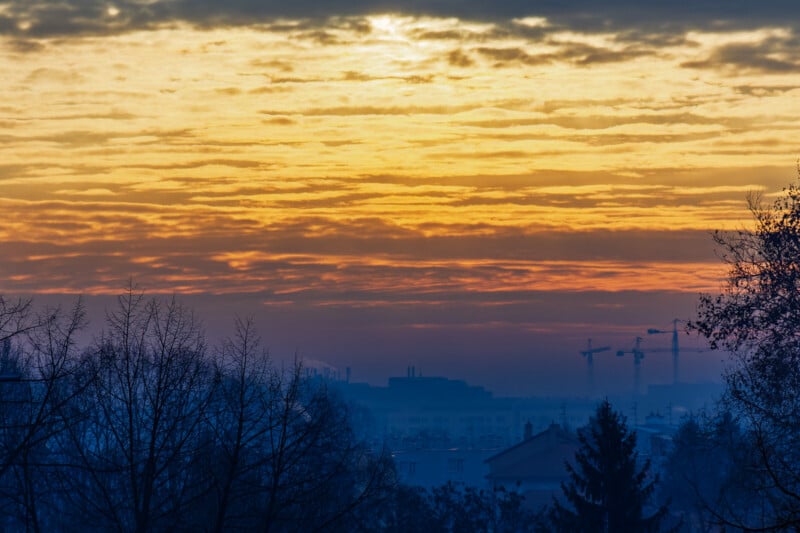 Horizonte de la ciudad al amanecer o al atardecer, con capas de nubes naranjas y amarillas sobre edificios industriales, grúas y árboles desnudos en primer plano.