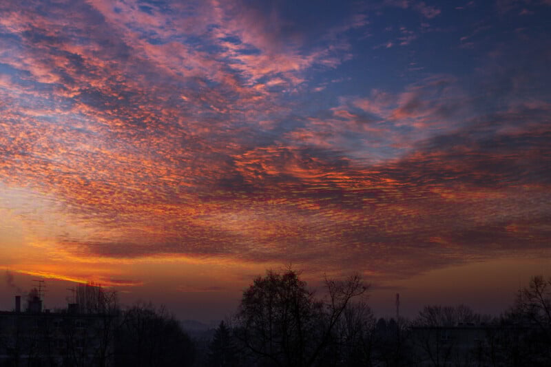 Un vibrante cielo al atardecer con nubes naranjas y moradas que salpican las siluetas de árboles y tejados, proyectando un cálido resplandor sobre el paisaje.