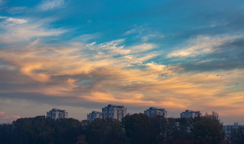 Un colorido cielo nocturno con nubes naranjas y azules sobre varios edificios de apartamentos de media altura, parcialmente oscurecido por una hilera de árboles en primer plano.