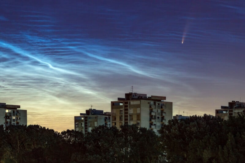 El complejo de apartamentos se alza contra un cielo crepuscular con nubes centelleantes y un cometa brillante o una estrella fugaz que pasa en diagonal sobre el horizonte; Los árboles se alinean en la parte inferior de la escena.