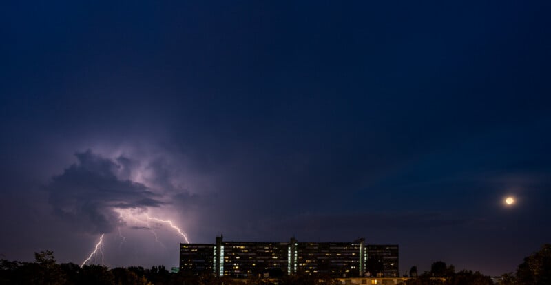 En el oscuro cielo nocturno, un relámpago brillante cayó sobre un gran edificio y las ventanas se iluminaron. La luna es visible en la esquina superior derecha y las siluetas de los árboles en primer plano.