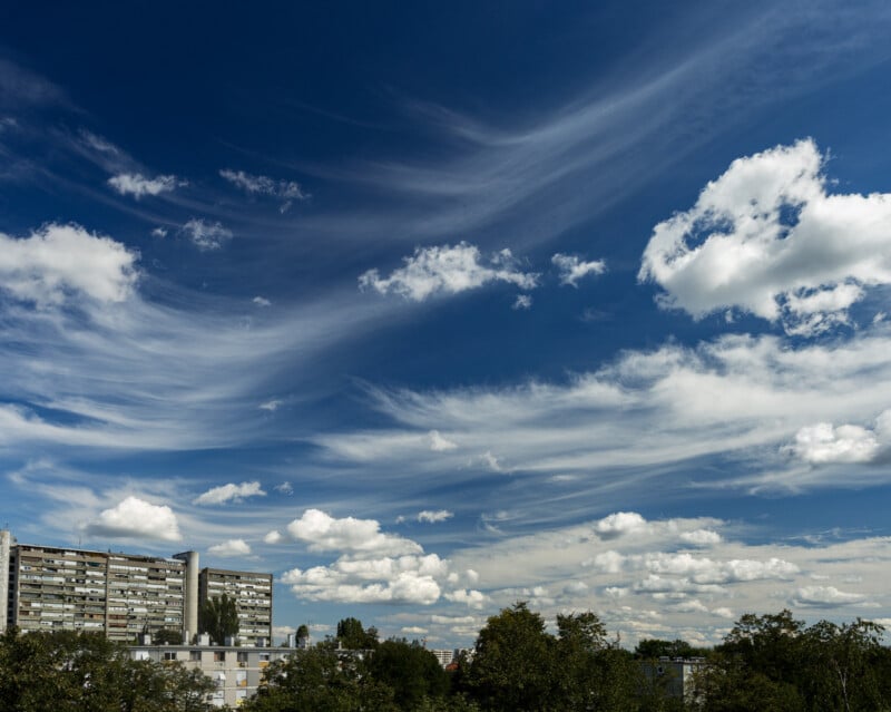 Edificios de apartamentos y árboles bajo un cielo azul brillante, salpicados de nubes blancas y jirones de cirros.