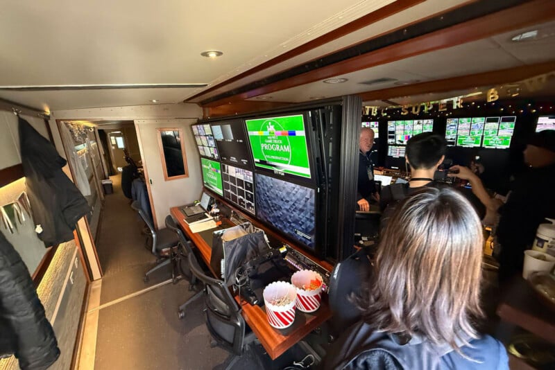 A production control room with large monitors displaying video feeds, equipment, and several people working. Popcorn tubs are on the desk, and the setting appears busy and focused.