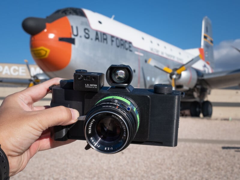 A hand holds a large, vintage-style camera with a visible lens and light meter in front of a parked U.S. Air Force aircraft under a clear blue sky.