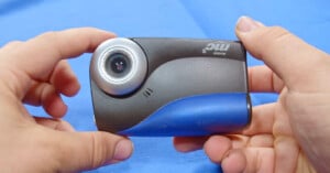 Close-up of two hands holding a compact black and blue camera with a circular lens on the left side, against a blue background.