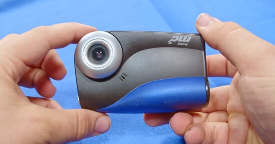 Close-up of two hands holding a compact black and blue camera with a circular lens on the left side, against a blue background.