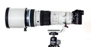 A white OM System telephoto camera lens attached to a black OM-1 Mark II camera is mounted on a tripod with a white background.