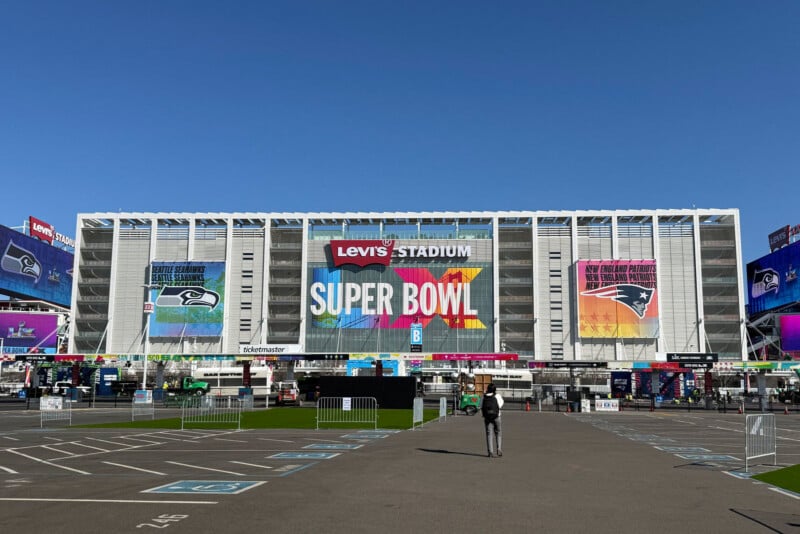 Levi's Stadium with large "SUPER BOWL" signage and team banners displayed on the exterior. One person walks toward the entrance in the mostly empty parking lot under a clear blue sky.