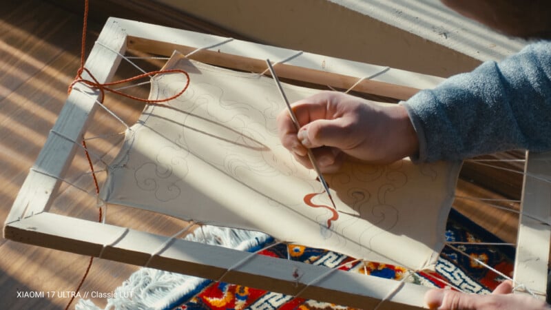 A person’s hand uses a brush to paint a red line on a detailed drawing secured to a wooden frame, with sunlight casting shadows and a colorful patterned rug visible below.