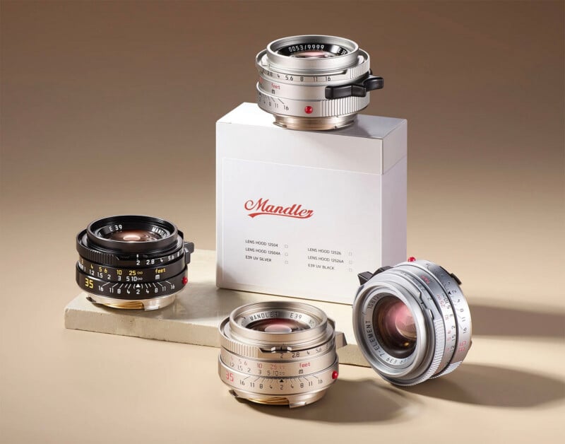 Four camera lenses are displayed on beige surfaces, with one placed on a white box labeled "Mandler." The lenses vary in silver and black finishes, and their focal lengths and aperture markings are visible.