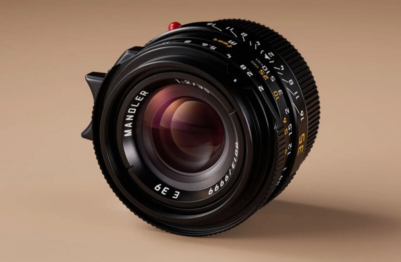 A close-up of a camera lens with the brand name "Manoler" and various numbers and markings visible, set against a beige background. The lens has a glossy finish and detailed focus and aperture rings.