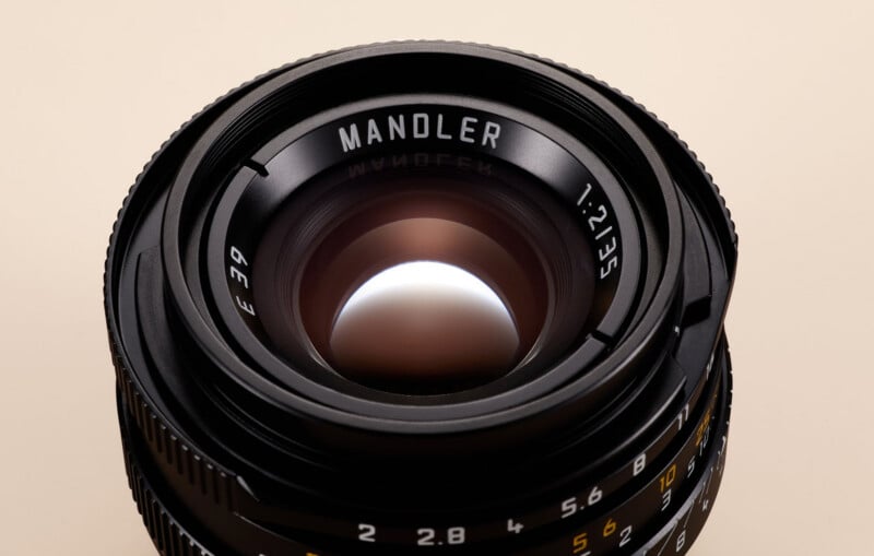 A close-up of a camera lens marked "MANDLER 1:2/35," showing the glass elements and aperture blades, with focus and aperture markings visible on the lens barrel against a beige background.